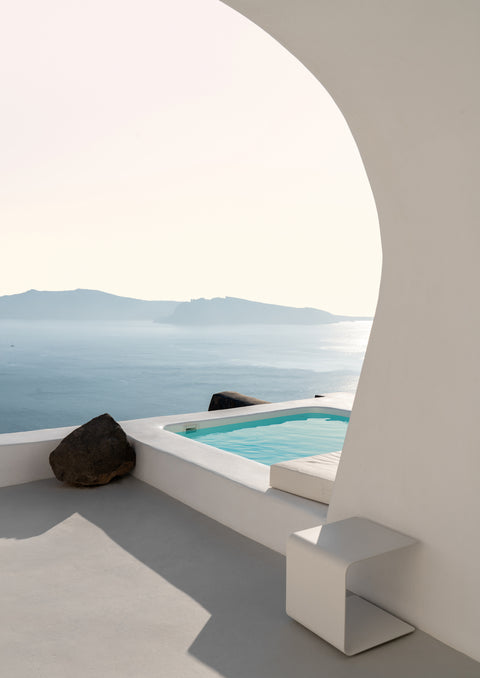 Minimalistic view of a swimming pool next to a white architectural structure, with a serene blue sea and distant mountains in the background.