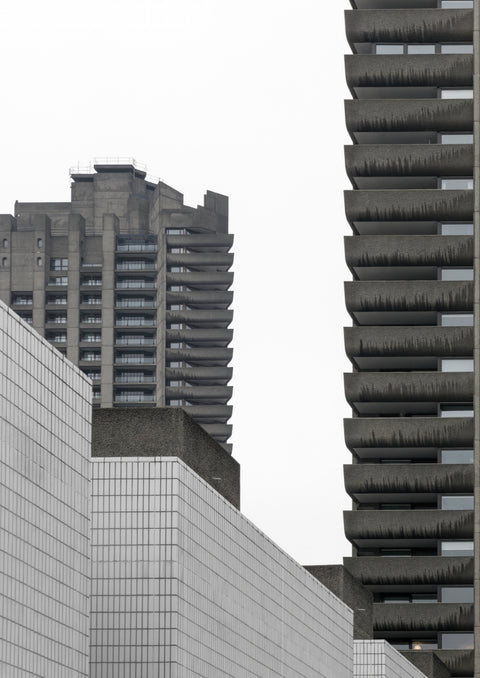 Two distinct high-rise buildings of the Barbican Estate captured from a street view, showcasing their brutalist architecture against a gray sky.